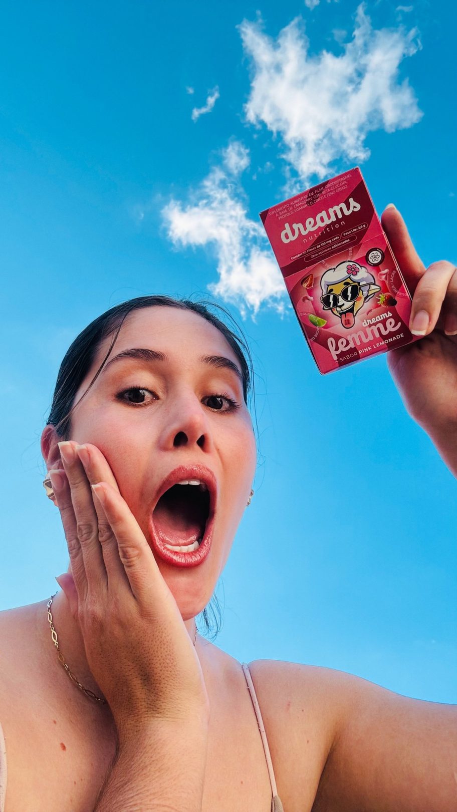 Laura Oliveira sorrindo animada segurando uma caixa de produto, com o céu azul ao fundo